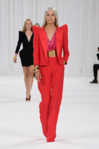 Older model in a bold red pantsuit with oversized shoulders, pink top, and gold belt walking a runway.