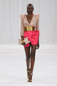Model walking a fashion show wearing a peach sheer blouse with pink high-waisted shorts, a wide gold belt, and gold-strap sandals, carrying a beige clutch edgewise.