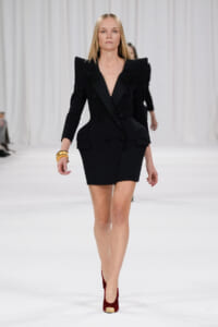 Female model walking a runway in a black double-breasted blazer dress with oversized shoulders and red pumps.