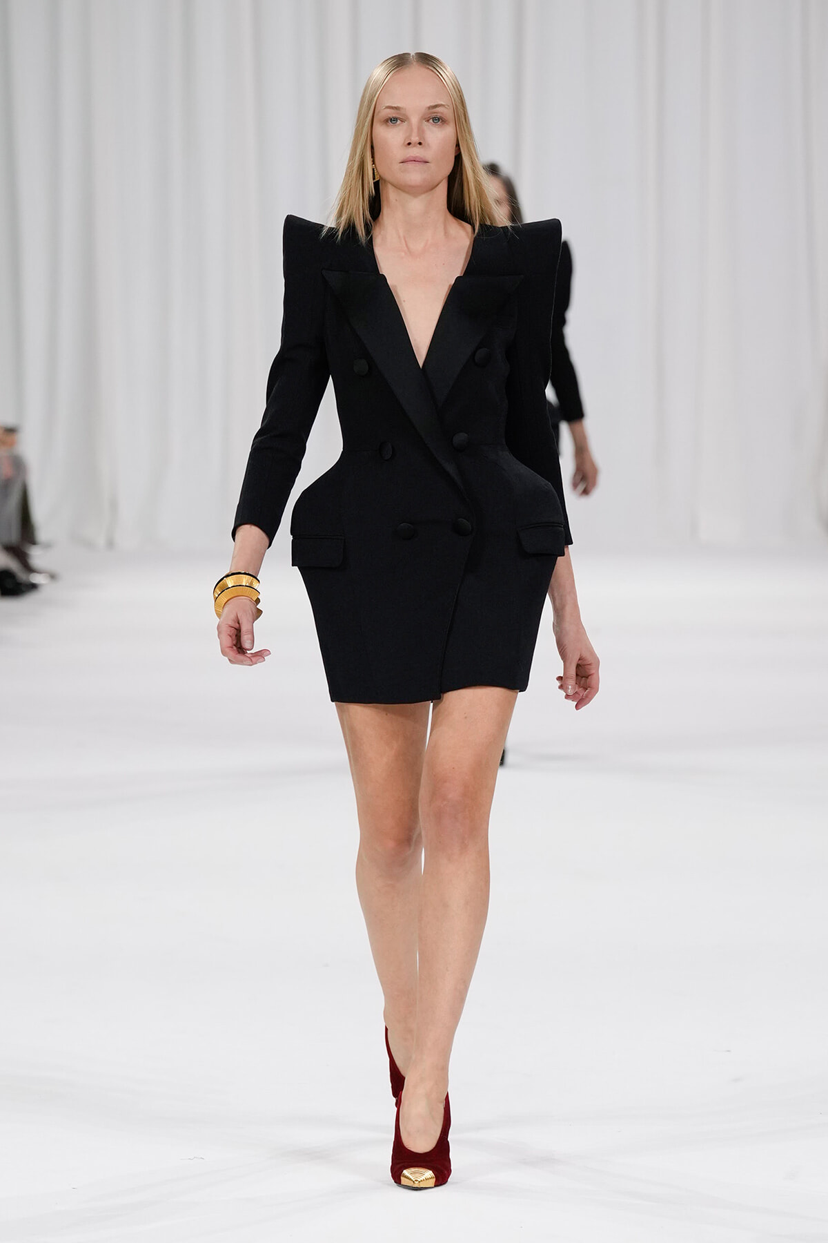 Female model walking a runway in a black double-breasted blazer dress with oversized shoulders and red pumps.