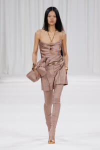 Model walks the runway in a dusty pink satin camisole with matching layered shorts and thigh-high boots, paired with gold jewelry and a pink clutch.