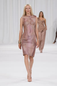 Model walks the runway in a sleeveless, mauve-toned fitted dress with a glossy, textured surface and matching heels.