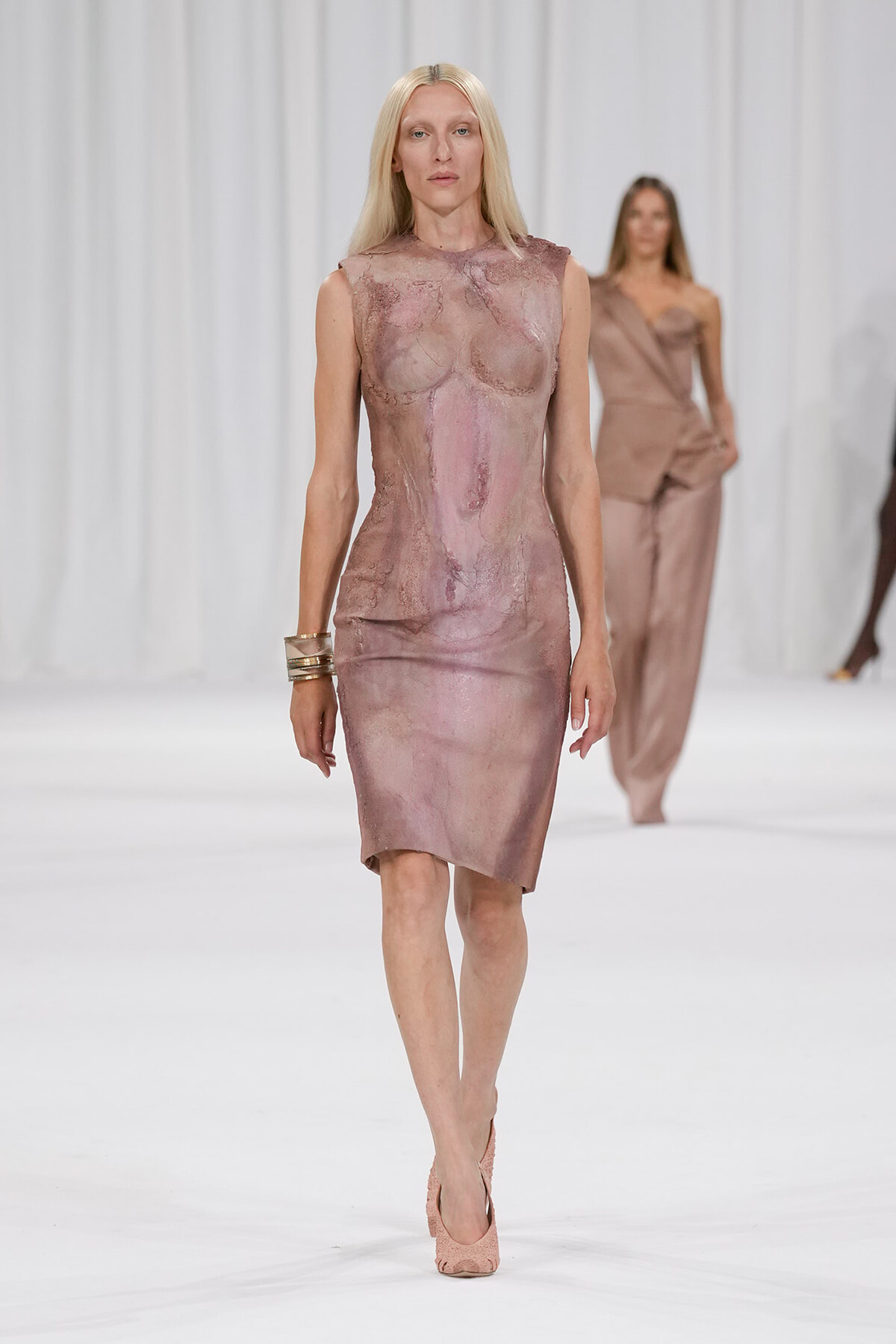 Model walks the runway in a sleeveless, mauve-toned fitted dress with a glossy, textured surface and matching heels.