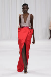 Runway model wearing a taupe plunging-neck top tucked into a bright red wrap skirt with a front slit and black strappy sandals.