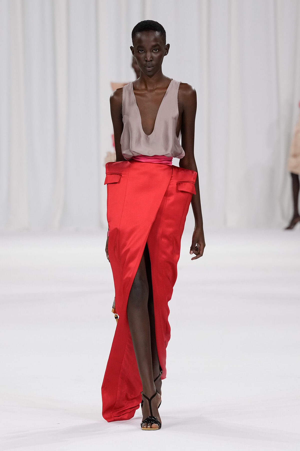 Runway model wearing a taupe plunging-neck top tucked into a bright red wrap skirt with a front slit and black strappy sandals.
