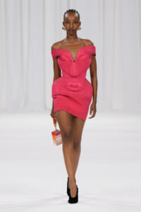 Model on a runway wearing a bright pink off‑the‑shoulder mini dress with a sculpted lip motif, black heels, braided hair, and a small multicolor box handbag.