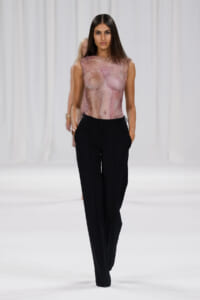 Model on a runway wearing a sheer pink marble-patterned sleeveless top and black wide-leg trousers on a white catwalk.