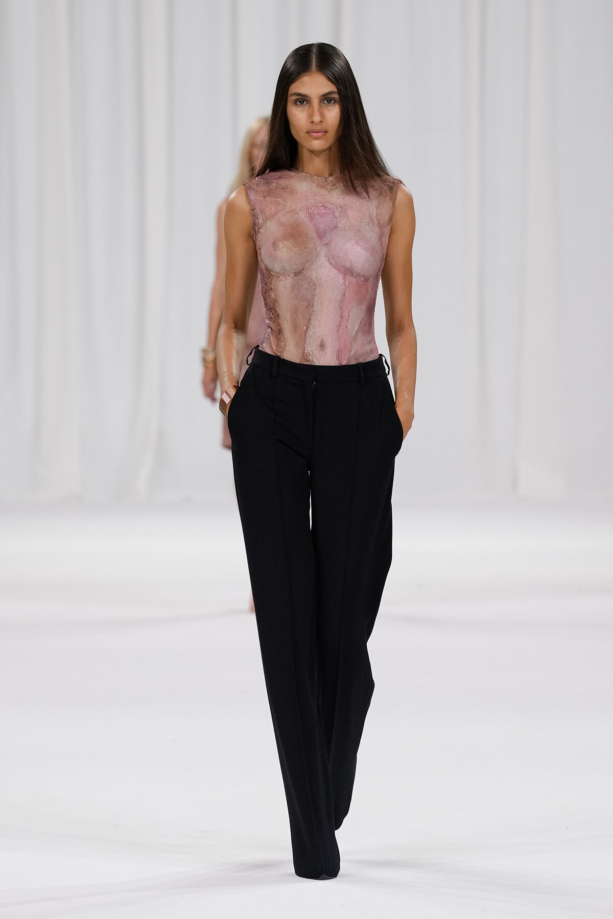 Model on a runway wearing a sheer pink marble-patterned sleeveless top and black wide-leg trousers on a white catwalk.