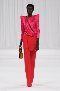 Model walks a white runway wearing a hot pink satin top with structured shoulders and high-waisted red cargo-style pants; she holds a small amber perfume bottle in her right hand.