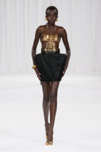 Model walking a runway in a metallic gold bandeau top and high-waisted black sculptural mini skirt with gold accessories.