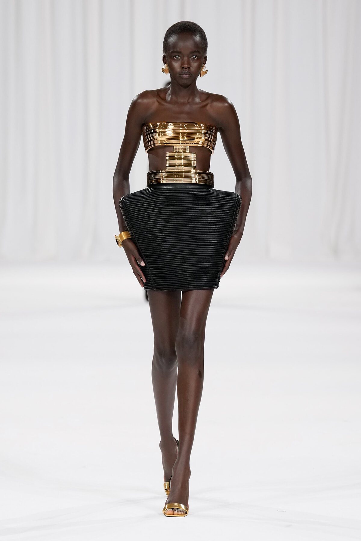 Model walking a runway in a metallic gold bandeau top and high-waisted black sculptural mini skirt with gold accessories.