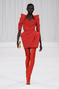 Runway model in a bold red sculpted dress with oversized shoulders and matching thigh-high boots, on a white catwalk."]} }{