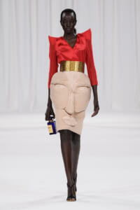 Runway model in a red satin V-neck blouse with padded shoulders, a wide gold belt, and a beige sculpted pencil skirt, carrying a blue bottle.