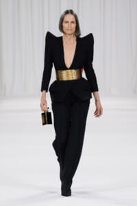 Runway look: black structured suit with dramatic shoulders, a wide gold belt, and a small black-and-gold handbag held at the side.