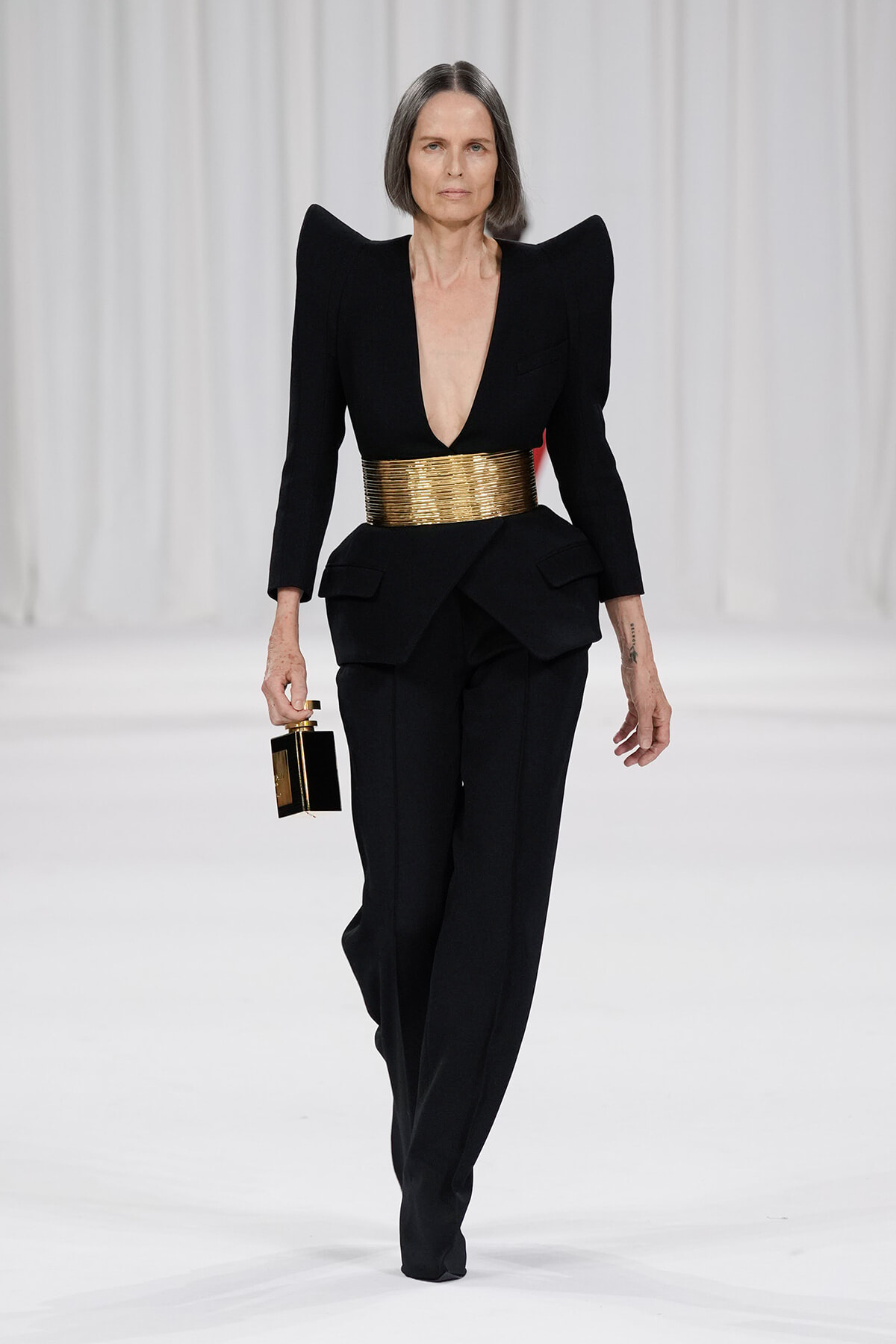 Runway look: black structured suit with dramatic shoulders, a wide gold belt, and a small black-and-gold handbag held at the side.