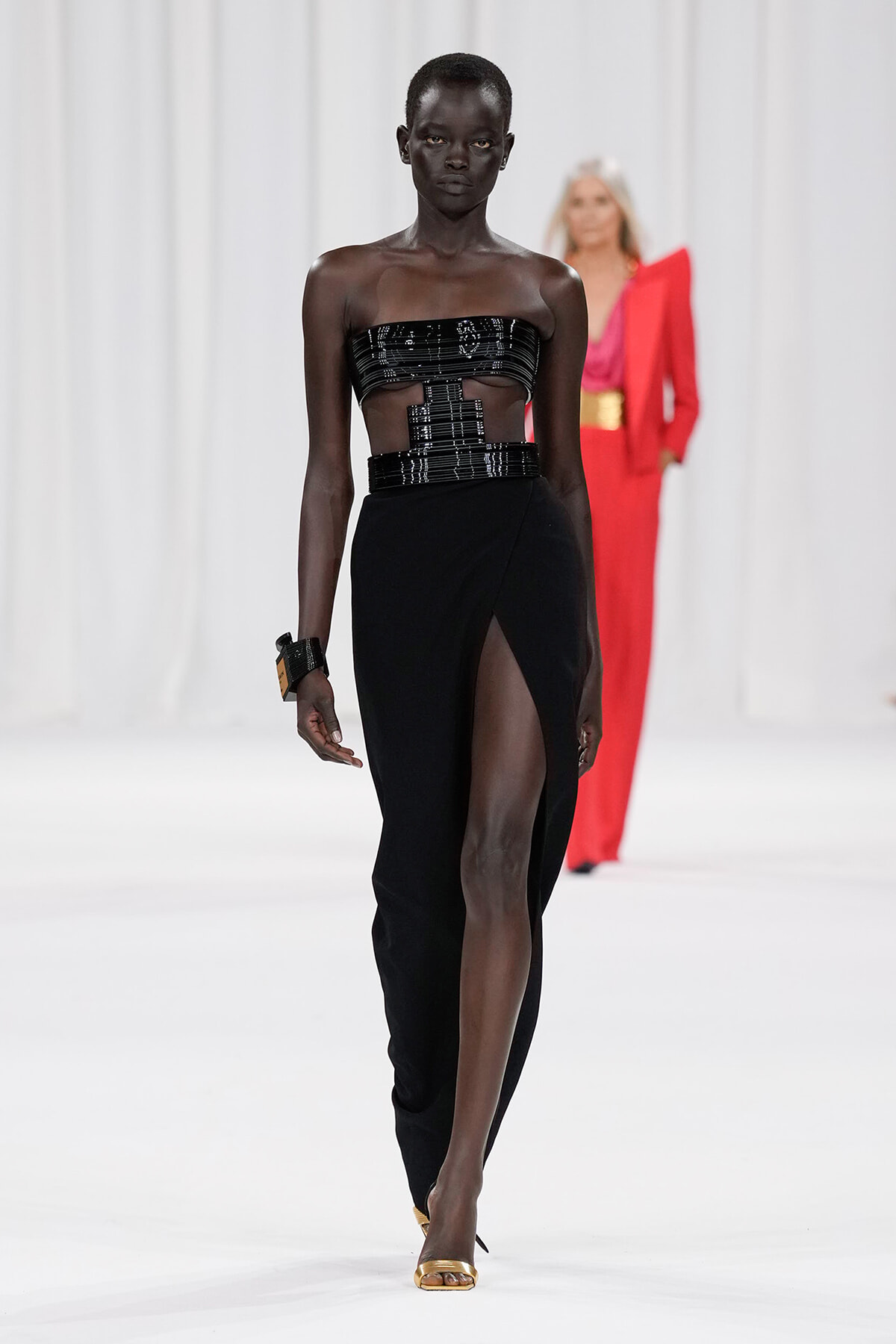 Runway model in a black strapless top with cutouts and a high-slit black skirt, walking the catwalk in gold heels.