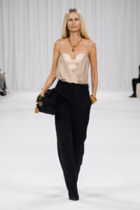 Model on a white runway wearing a beige draped camisole with black high-waisted trousers, gold jewelry, and a black handbag.