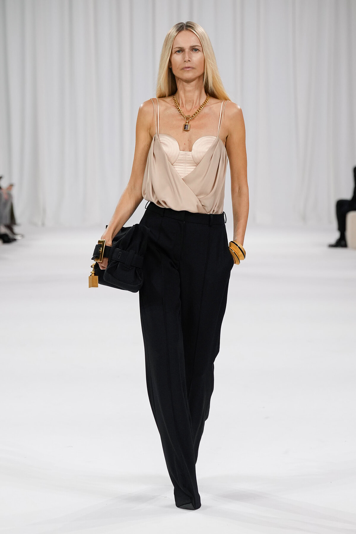 Model on a white runway wearing a beige draped camisole with black high-waisted trousers, gold jewelry, and a black handbag.