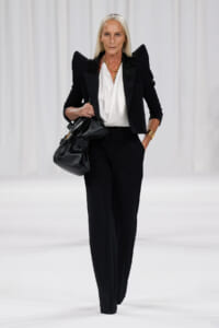 Runway model in a tailored black suit with a white draped blouse, gold jewelry, and a black handbag.