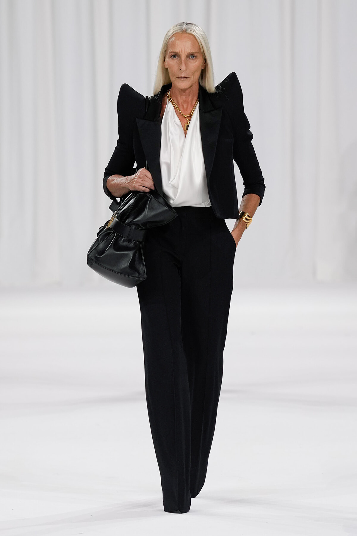 Runway model in a tailored black suit with a white draped blouse, gold jewelry, and a black handbag.