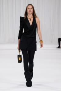 Model walks a runway in an all-black ensemble: draped one-shoulder top with a structured blazer and tailored pants, gold jewelry, and a black rectangular handbag with gold accents.