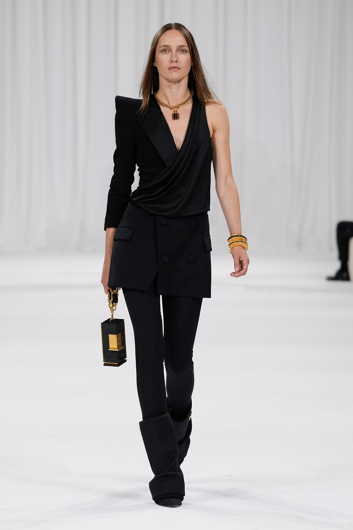 Model walks a runway in an all-black ensemble: draped one-shoulder top with a structured blazer and tailored pants, gold jewelry, and a black rectangular handbag with gold accents.
