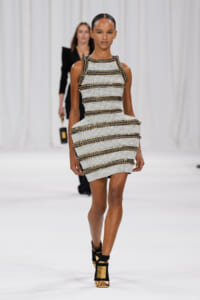 Model walks the runway in a white textured mini dress with gold chain trim and black-gold ankle-strap sandals.