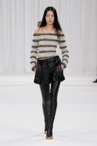 Model walking a runway in an off‑the‑shoulder knit top with white and black stripes and gold trim, paired with black leather shorts and thigh‑high boots.