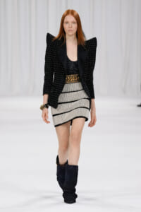 Model walking the runway in a black studded blazer with exaggerated shoulders, a black top, a striped mini skirt with a gold belt, and tall black boots.