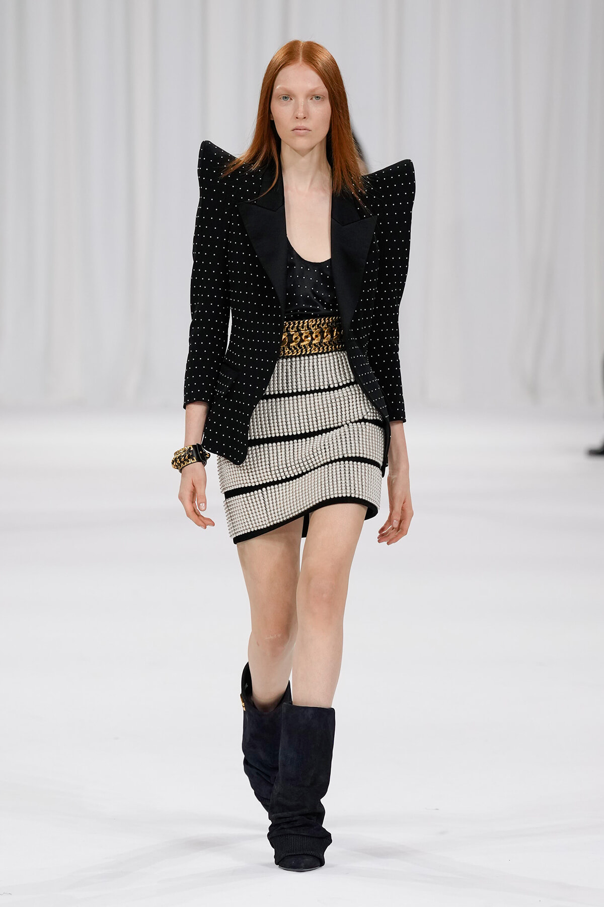Model walking the runway in a black studded blazer with exaggerated shoulders, a black top, a striped mini skirt with a gold belt, and tall black boots.