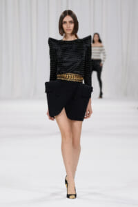 Model walking runway in a black studded top with dramatic shoulders and an asymmetric black skirt with a gold chain belt, black heels with gold toe accents.