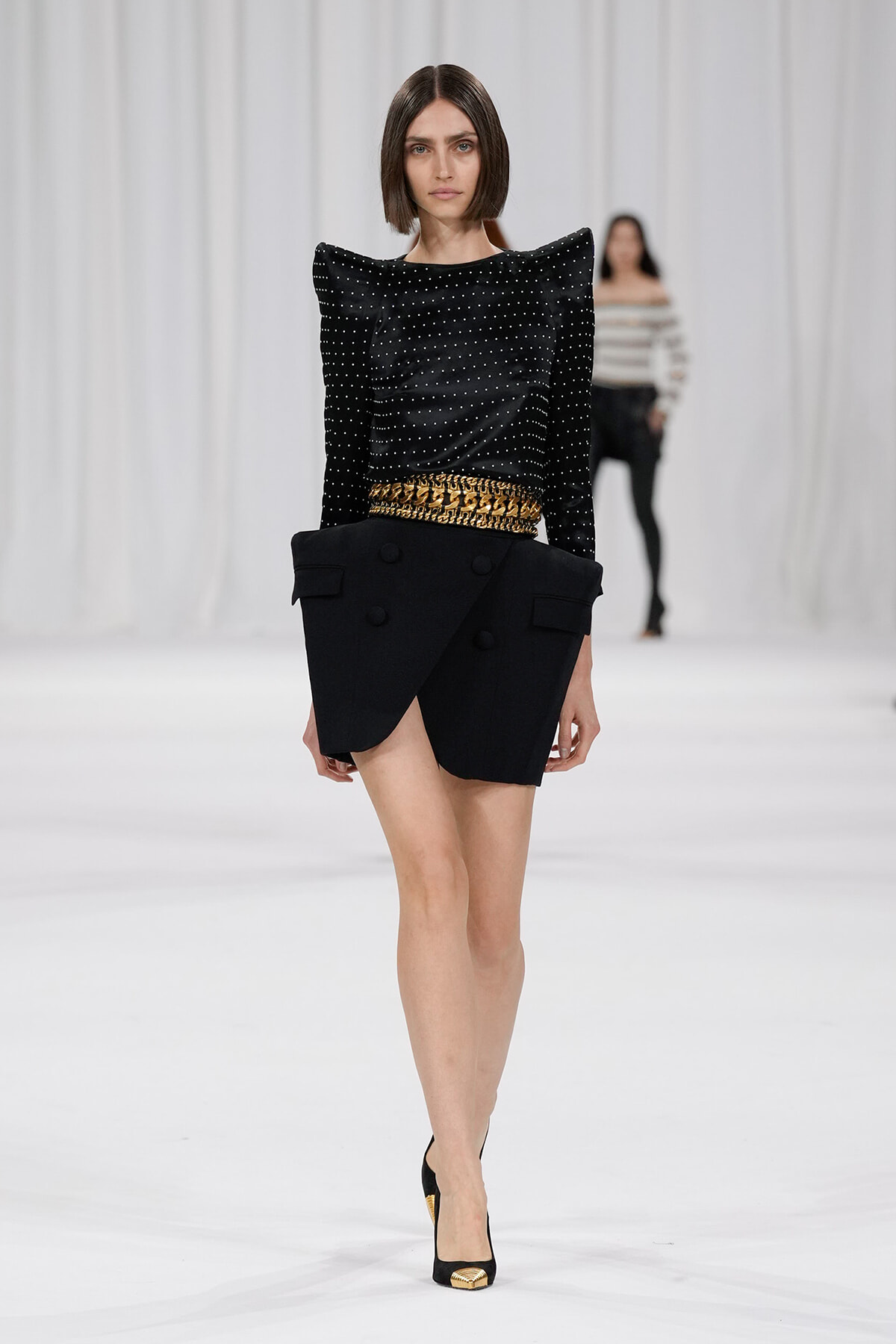 Model walking runway in a black studded top with dramatic shoulders and an asymmetric black skirt with a gold chain belt, black heels with gold toe accents.