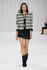 Model walking runway in a black-and-white studded jacket, black mini skirt, and slouchy black boots.