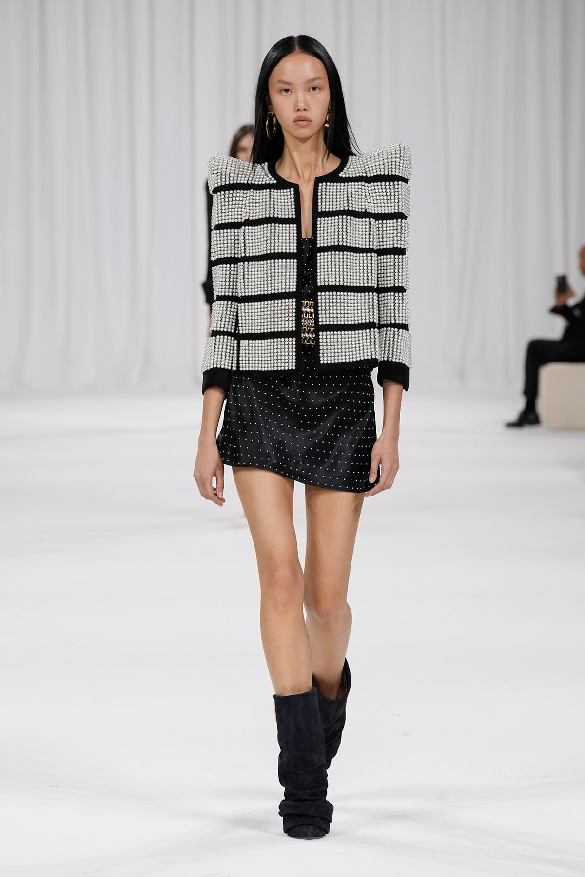 Model walking runway in a black-and-white studded jacket, black mini skirt, and slouchy black boots.