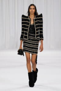 Model walking a runway in a black sequined striped suit with white trim and matching jacket, paired with black boots and a small handbag.