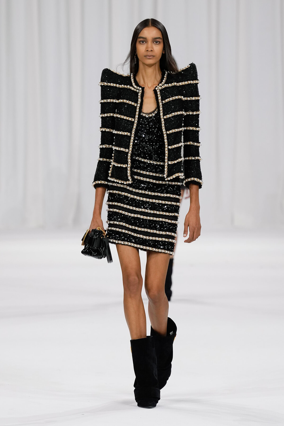 Model walking a runway in a black sequined striped suit with white trim and matching jacket, paired with black boots and a small handbag.