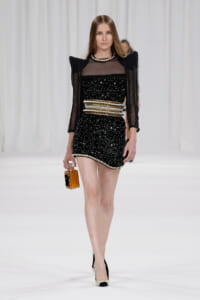 Model walking the runway in a black sequined mini dress with sheer sleeves and bold gold belt, holding a small perfume bottle.