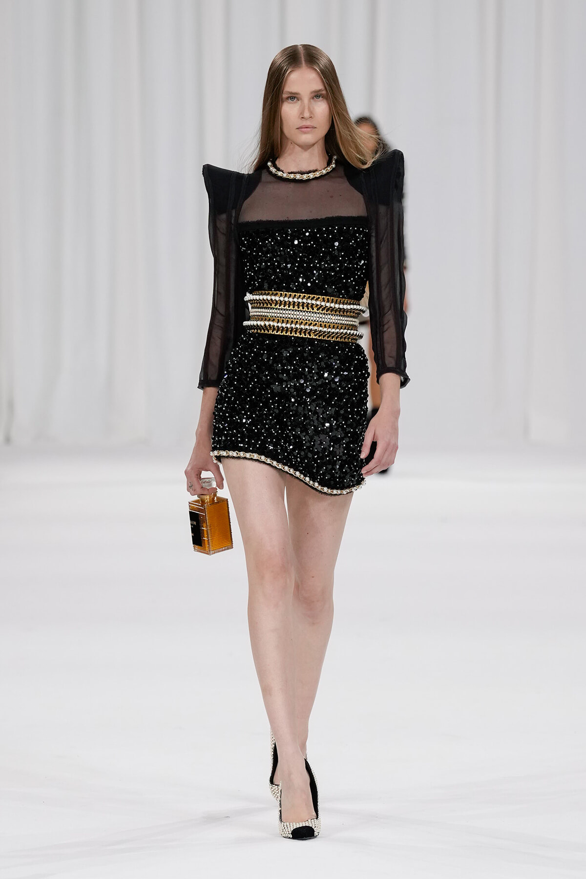 Model walking the runway in a black sequined mini dress with sheer sleeves and bold gold belt, holding a small perfume bottle.