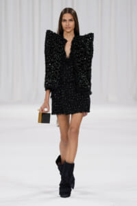 Model walking a runway in a black sequin ensemble with oversized shoulders, holding a small black-and-wood rectangular clutch, and wearing black boots.