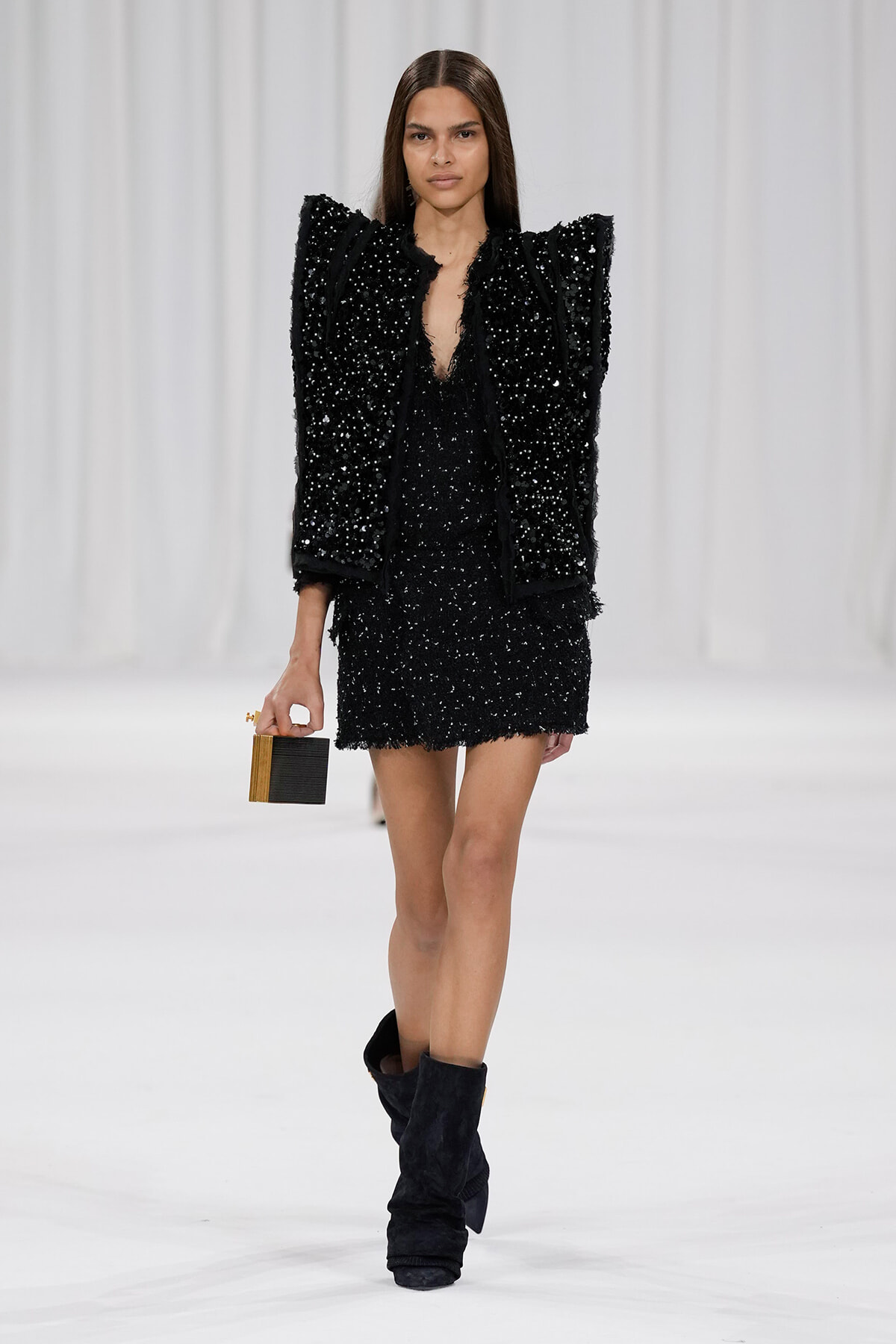 Model walking a runway in a black sequin ensemble with oversized shoulders, holding a small black-and-wood rectangular clutch, and wearing black boots.