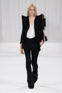 Fashion model on a white runway wearing a black textured blazer, white blouse, and black pants with tall boots.