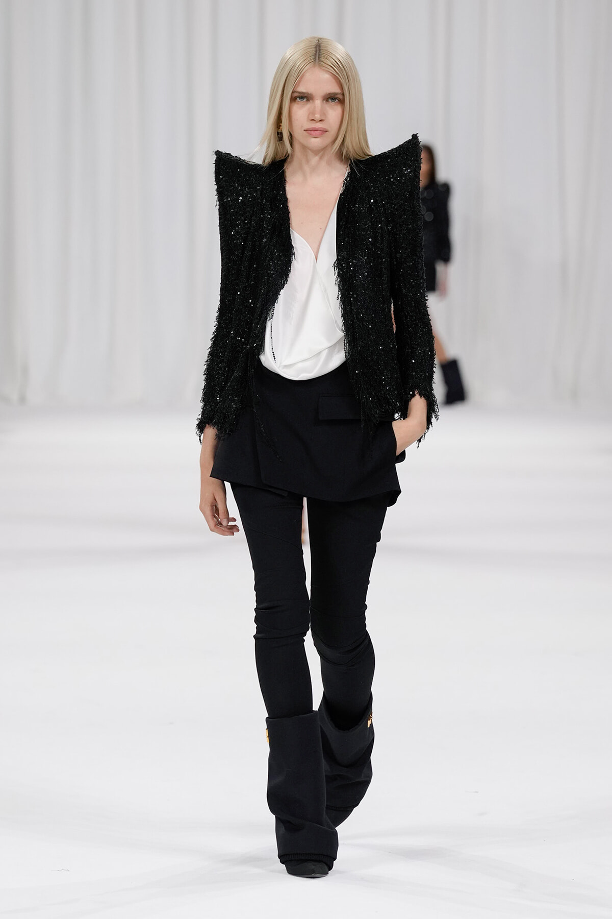 Fashion model on a white runway wearing a black textured blazer, white blouse, and black pants with tall boots.