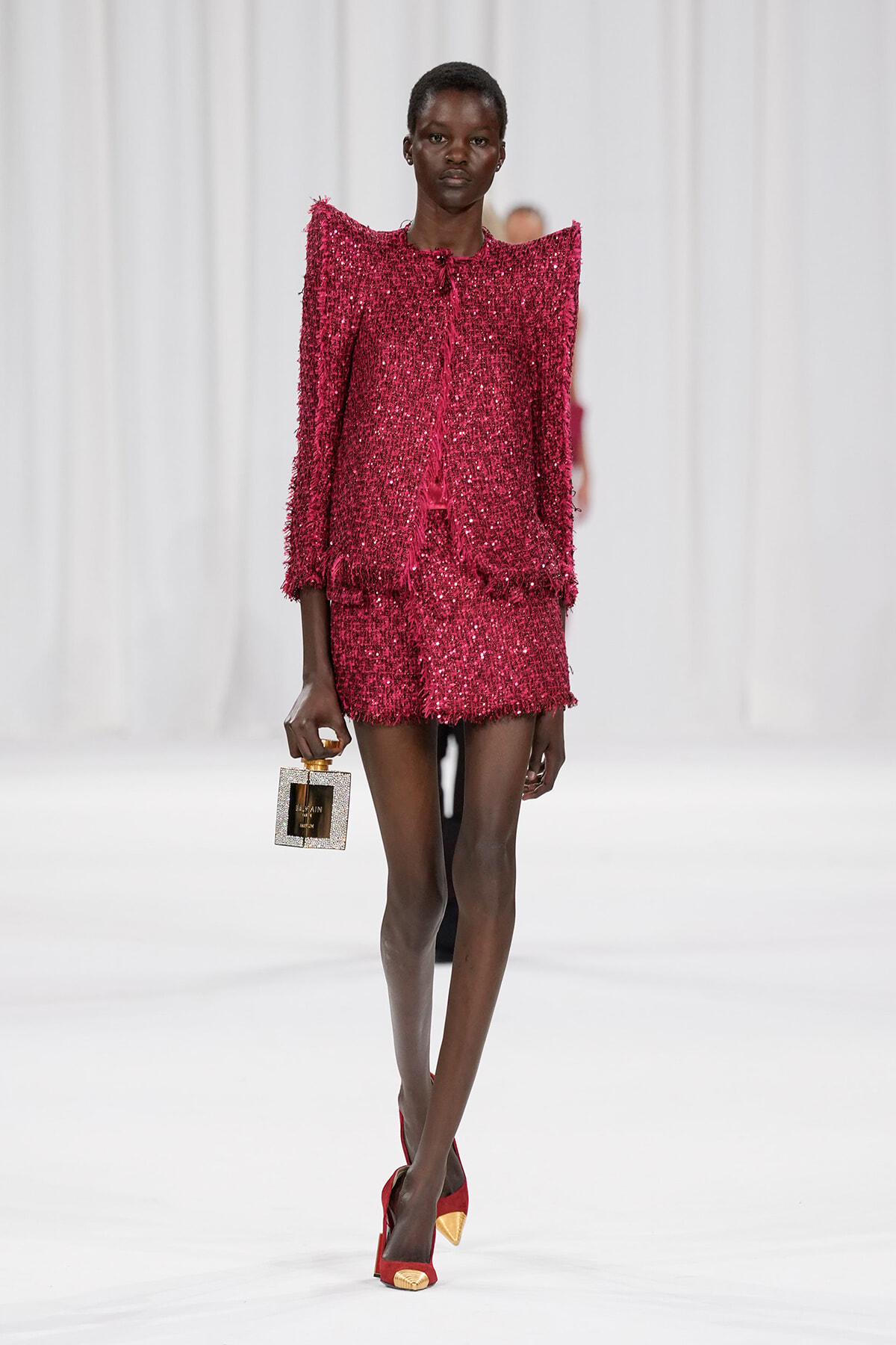 Model on a runway wearing a glittery pink fringe suit with a matching mini skirt, carrying a sparkle square handbag and red heels.