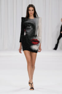 Model walking the runway in a sparkling two-tone mini dress with a large grayscale portrait and bright red lips on the front.