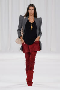 Model on runway wearing a gray structured blazer over a black top, burgundy suede thigh-high boots, and a gold pendant necklace.