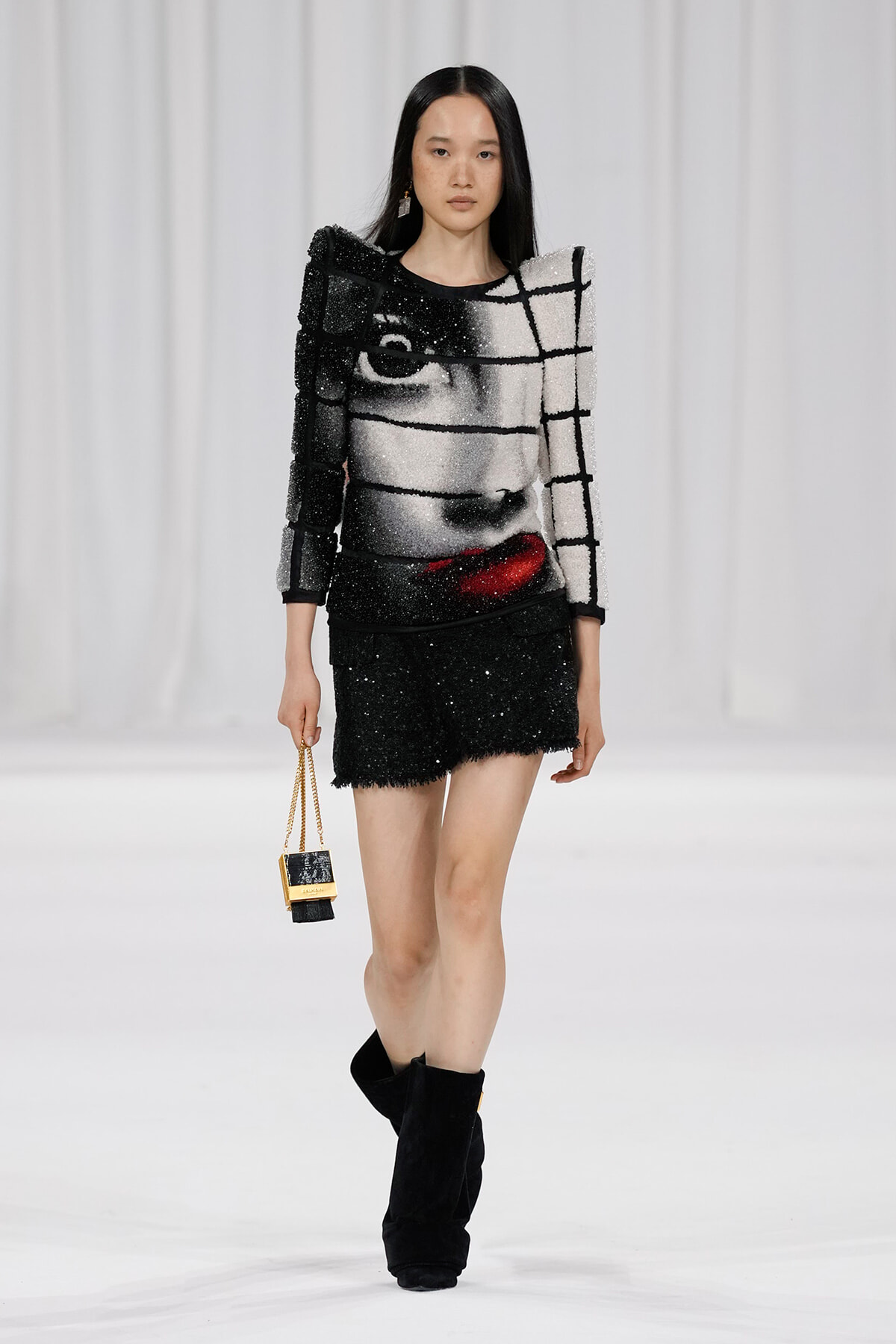 Model on a fashion runway wearing a black-and-white grid-pattern sequined top and matching black mini skirt, with black boots and a small gold-chain handbag.