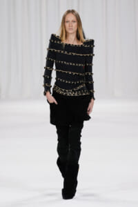 Model walking runway in black outfit with stacked gold chain bands across the torso and arms