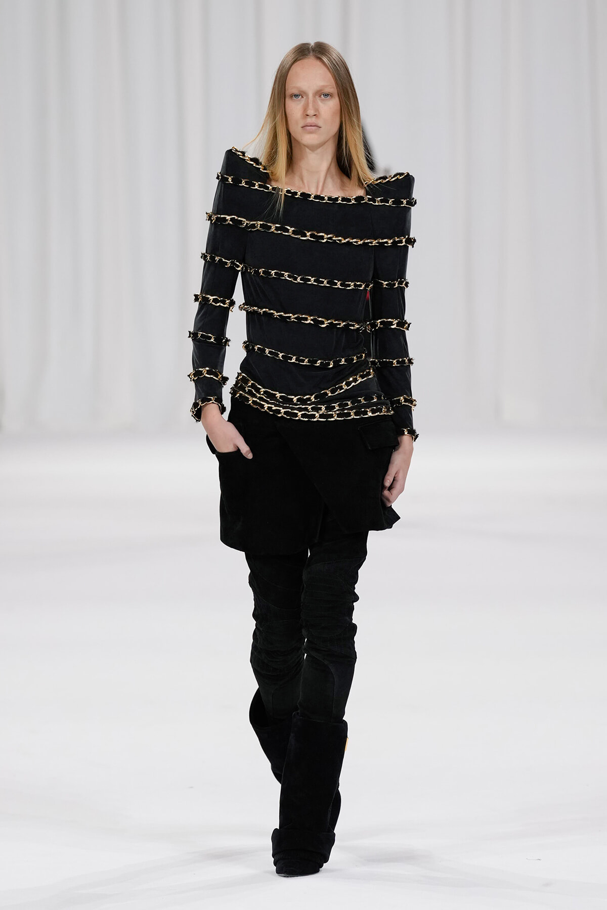 Model walking runway in black outfit with stacked gold chain bands across the torso and arms