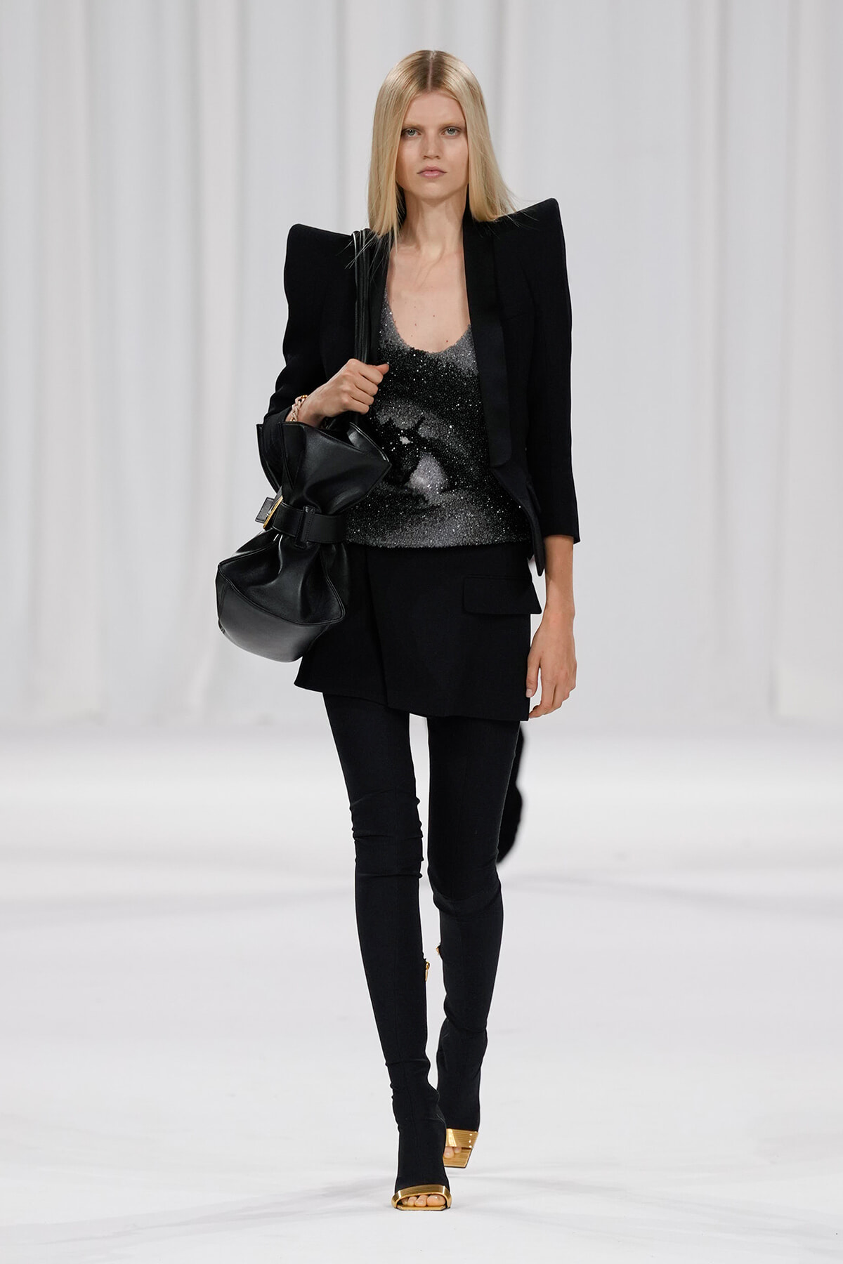 Female model walking runway in a black ensemble with oversized shoulder blazer, glittery top, and black leggings/skirt, carrying a black handbag.