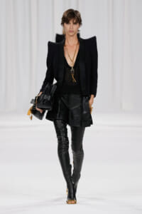 Fashion model on a runway in an all-black, high-fashion outfit with a structured blazer, leather shorts, and thigh-high boots, accessorized with gold jewelry and a bag.